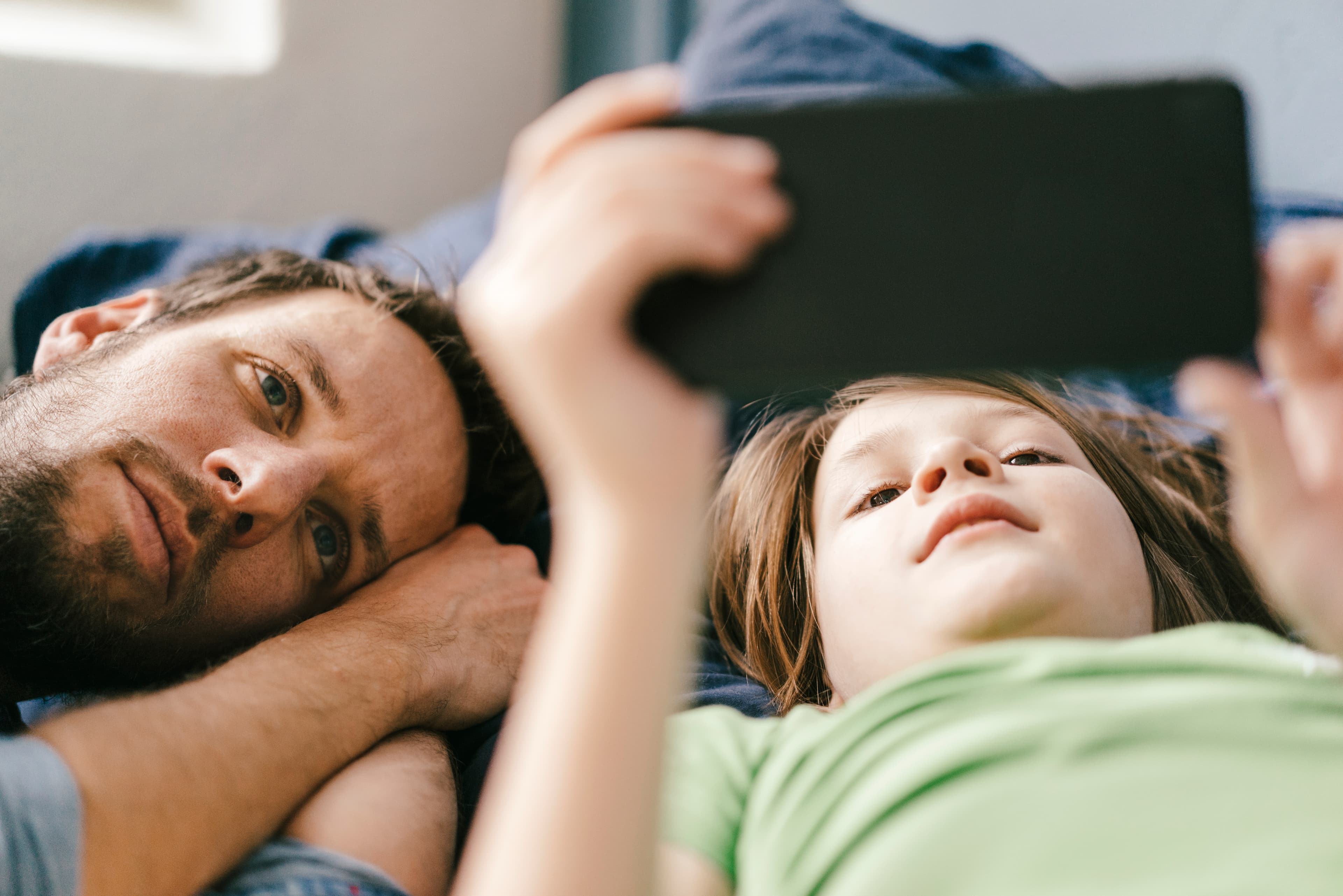 A father and his young child lying together, both watching something on a phone held above them in soft daylight.