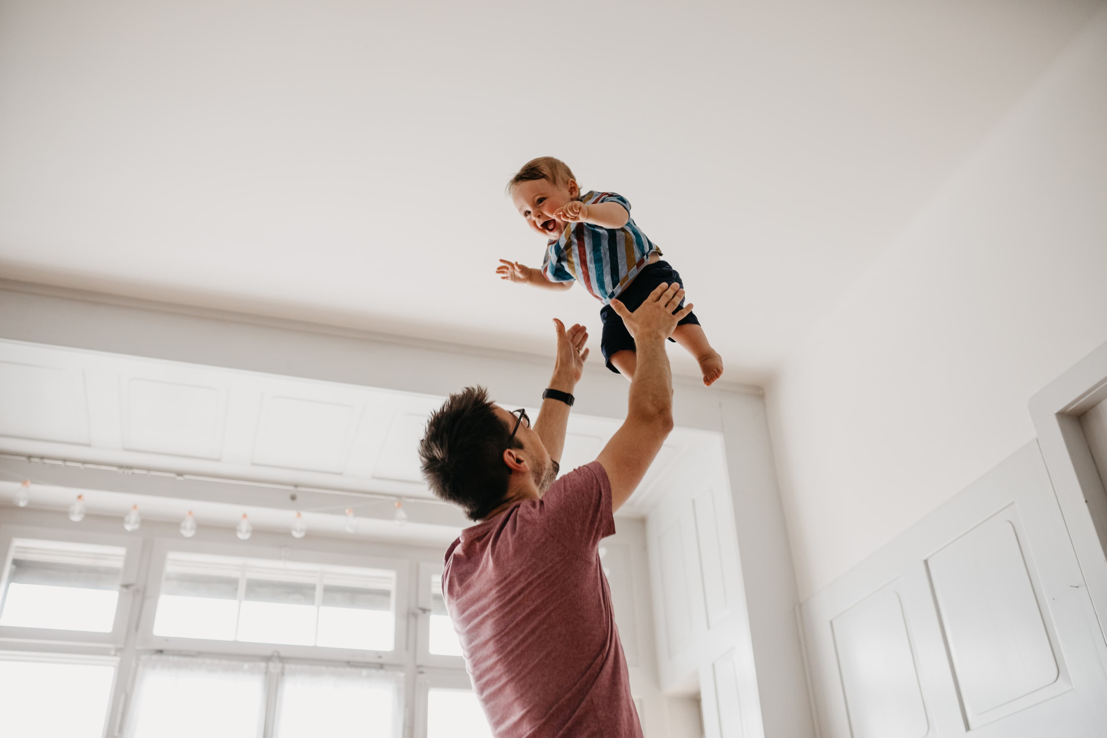 A father tossing his laughing toddler into the air in a bright, sunlit room.