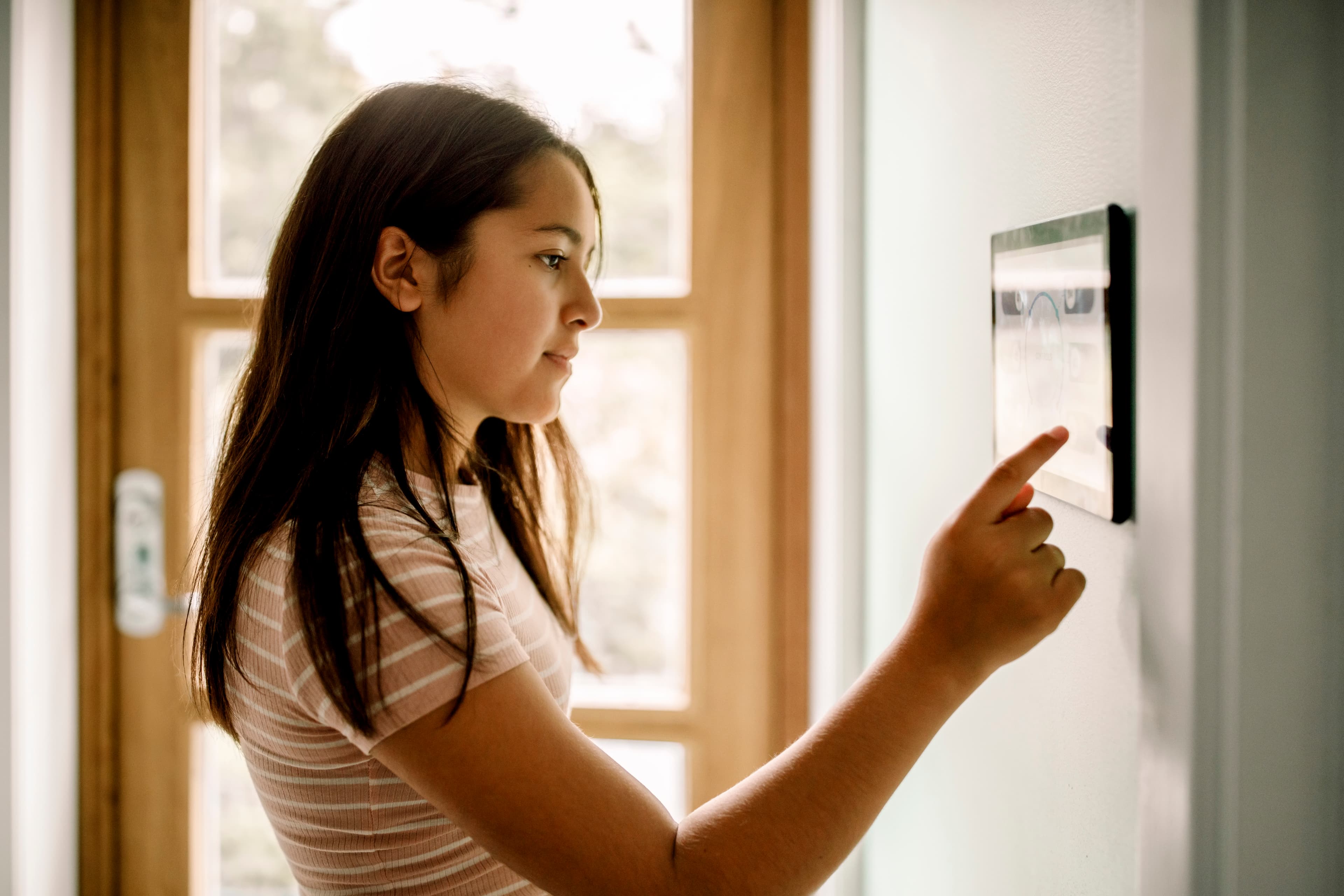 A teenager interacting with a wall-mounted home-energy display in a sunlit window bay.